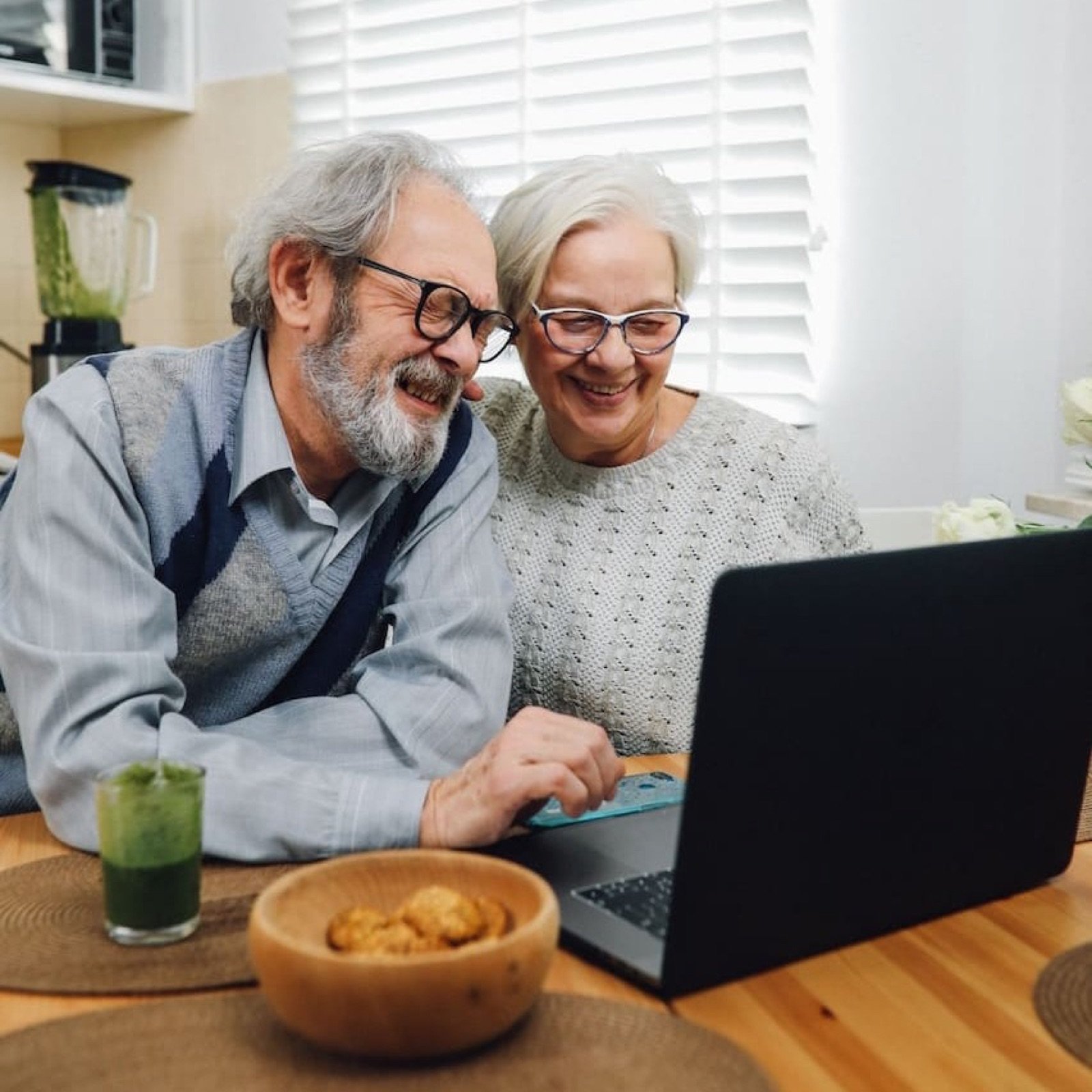 An older couple smiling at a laptop at their kitchen table